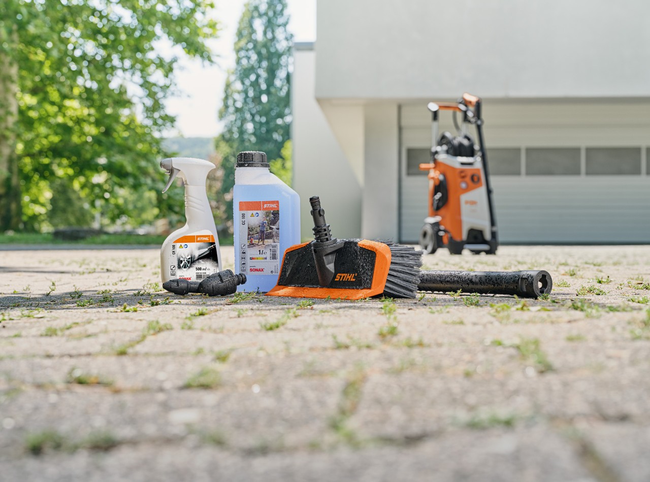  Środek do czyszczenia felg STIHL, środek do czyszczenia pojazdów i szczotka myjąca leżące na podłodze przed białym garażem. W tle myjka wysokociśnieniowa STIHL