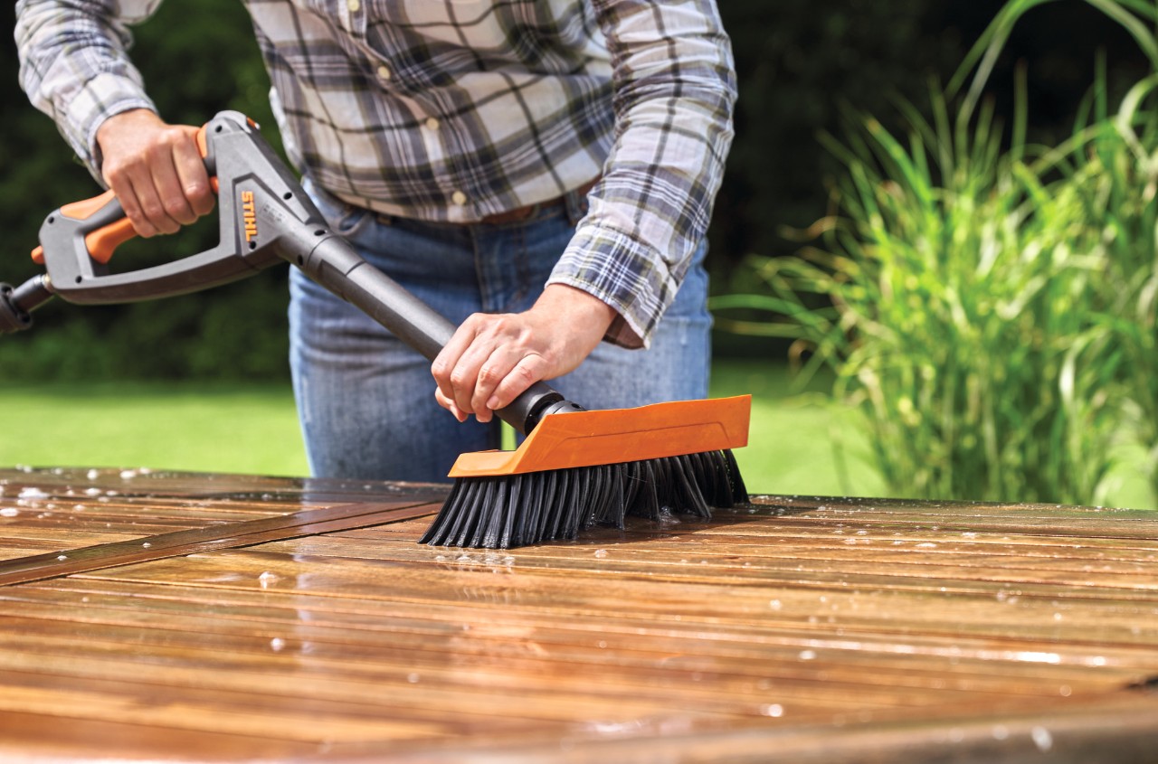 Kobieta czyści drewniany stół ogrodowy myjką wysokociśnieniową STIHL RE 130 plus z założoną odpowiednią szczotką do mycia