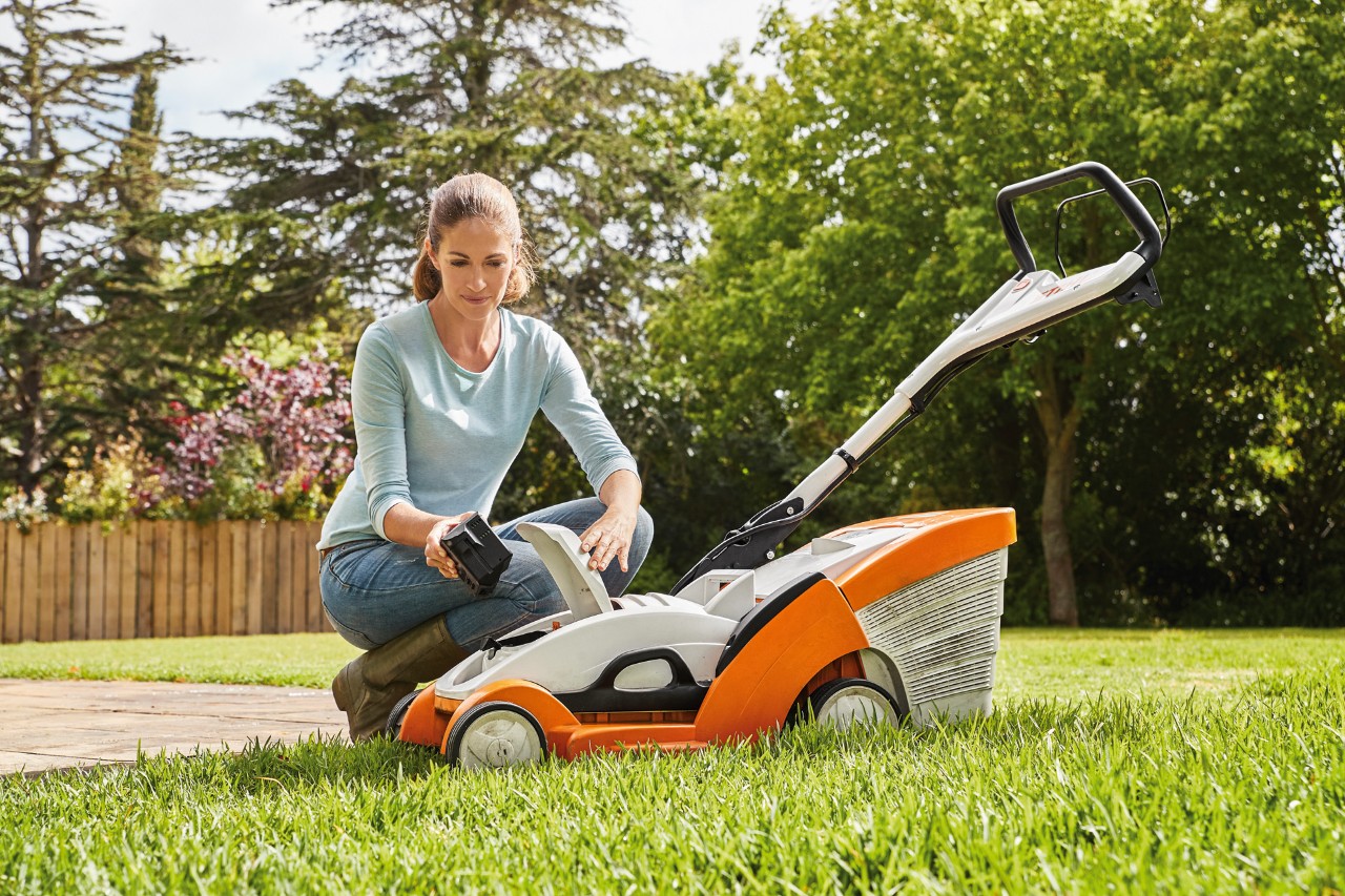 Kobieta klęczy obok kosiarki akumulatorowej STIHL RMA 339 C i wyjmuje akumulator