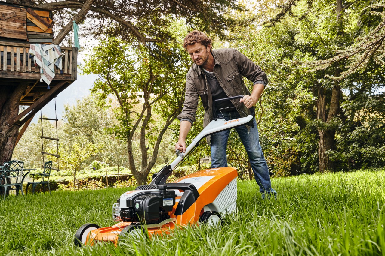 Mężczyzna na łące ogrodowej ciągnie za linkę rozrusznika kosiarki benzynowej STIHL RM 448 TC, aby ją uruchomić