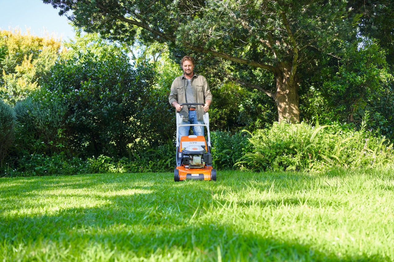 Mężczyzna kosi trawnik w półcieniu kosiarką STIHL, w tle krzewy i drzewa