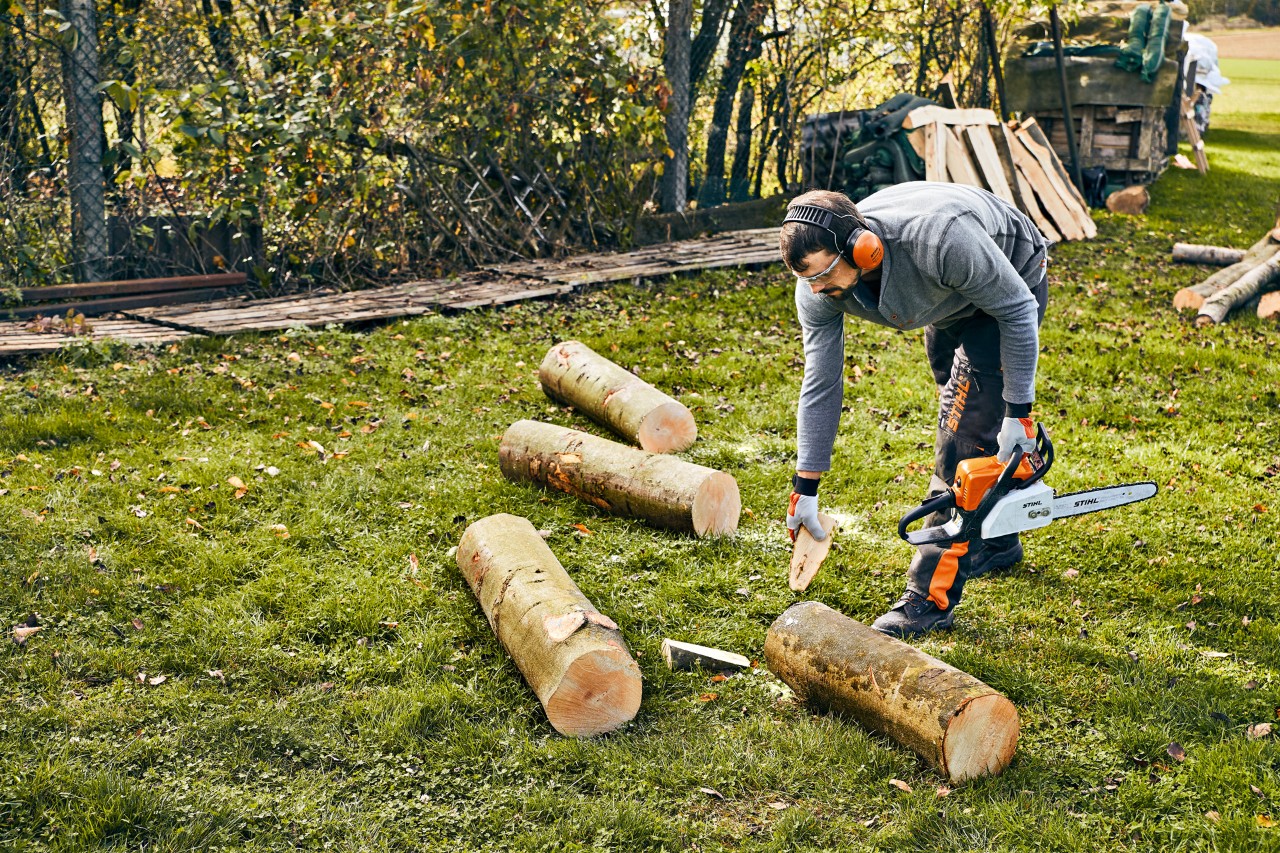 Metrowa kłoda jest zabezpieczana klinami przez osobę ubraną w środki ochrony indywidualnej i trzymającą spalinową piłę łańcuchową STIHL MS 180.
