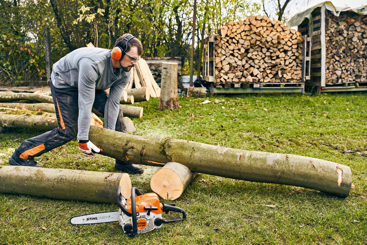 Pień drzewa zostaje ponownie umieszczony na stojaku do cięcia, a spalinowa piła łańcuchowa STIHL MS 180 stoi z przodu na ziemi.