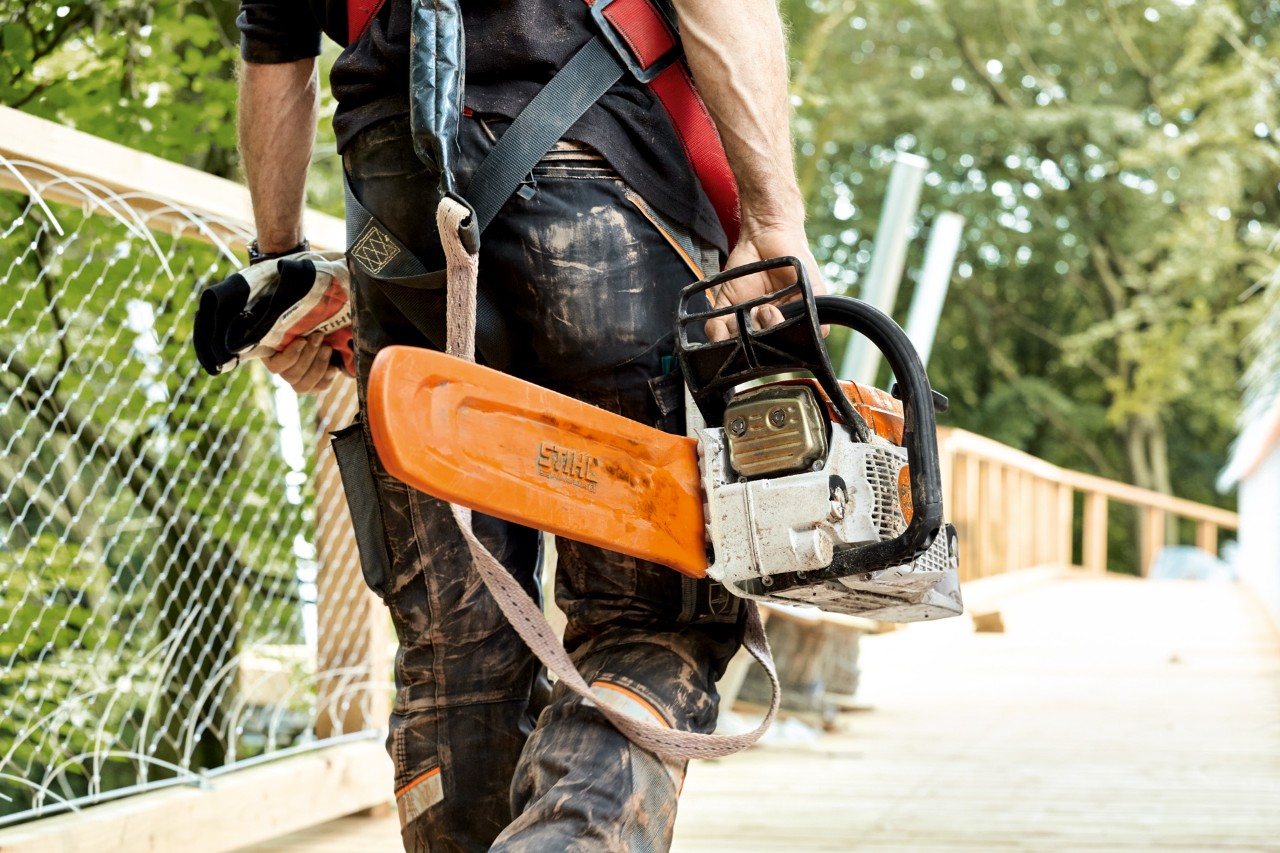 Mężczyzna w odzieży ochronnej niesie benzynową pilarkę łańcuchową STIHL MS 261 z osłoną łańcucha.