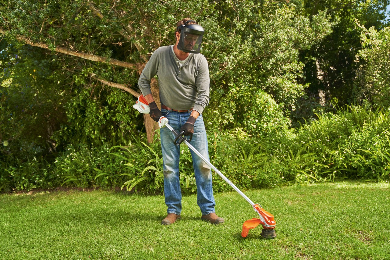 Mężczyzna z osłoną twarzy kosi trawnik w ogrodzie z akumulatorową kosą mechaniczną STIHL FSA 60