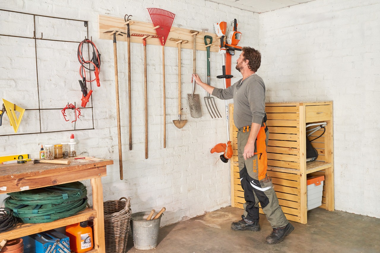Mężczyzna w spodniach roboczych STIHL wiesza łopatę na samodzielnie zbudowanym uchwycie na narzędzia ogrodowe obok kosy STIHL 