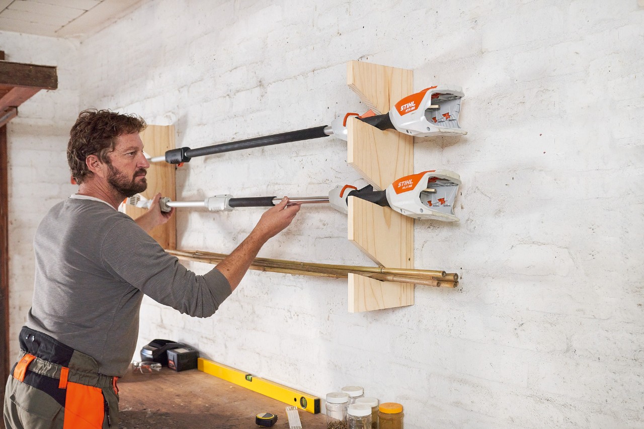 Mężczyzna zawiesza kosę mechaniczną STIHL na samodzielnie wykonanym uchwycie na narzędzia ogrodowe, przymocowanym do ściany 