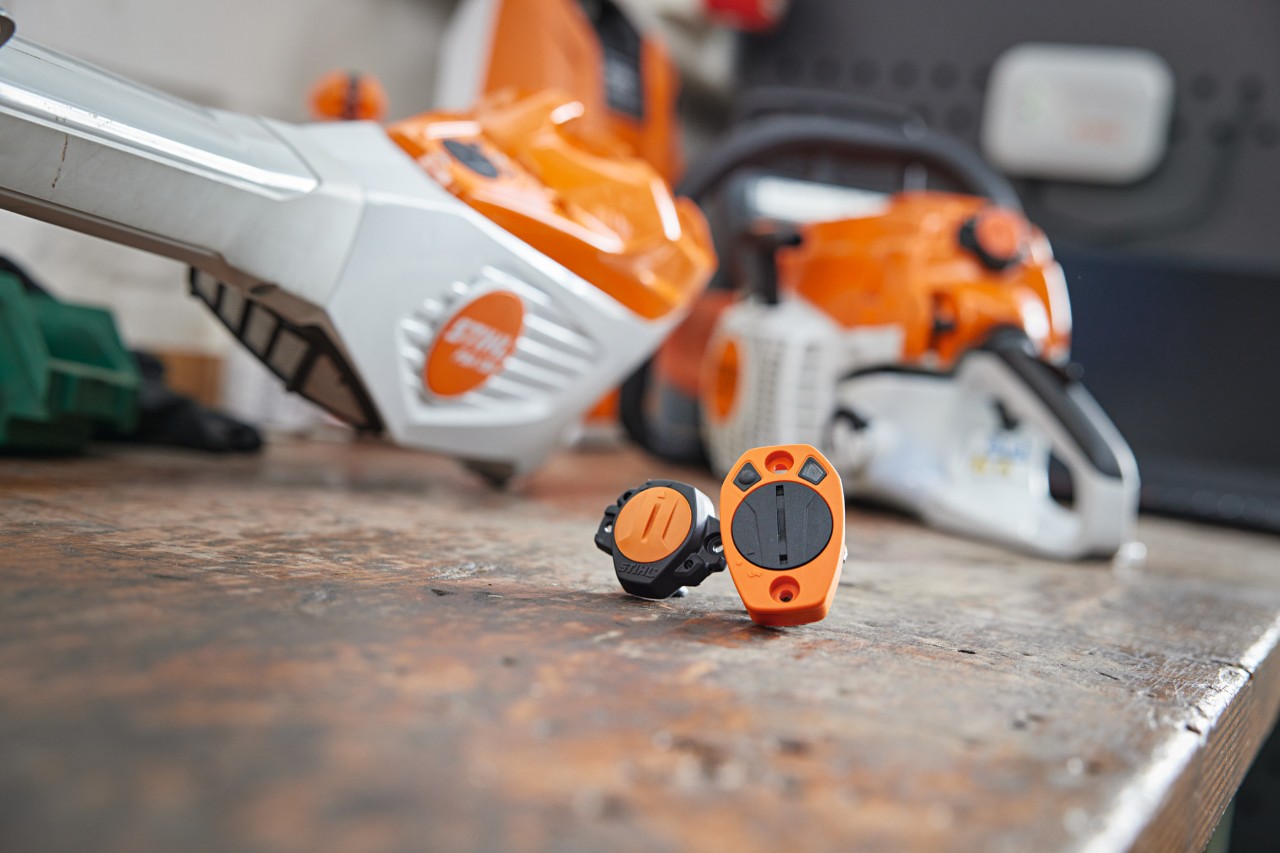 Złącza STIHL Smart Connector i STIHL Smart Connector 2 A leżą na stole warsztatowym. W tle widać rozmyte różne urządzenia STIHL.