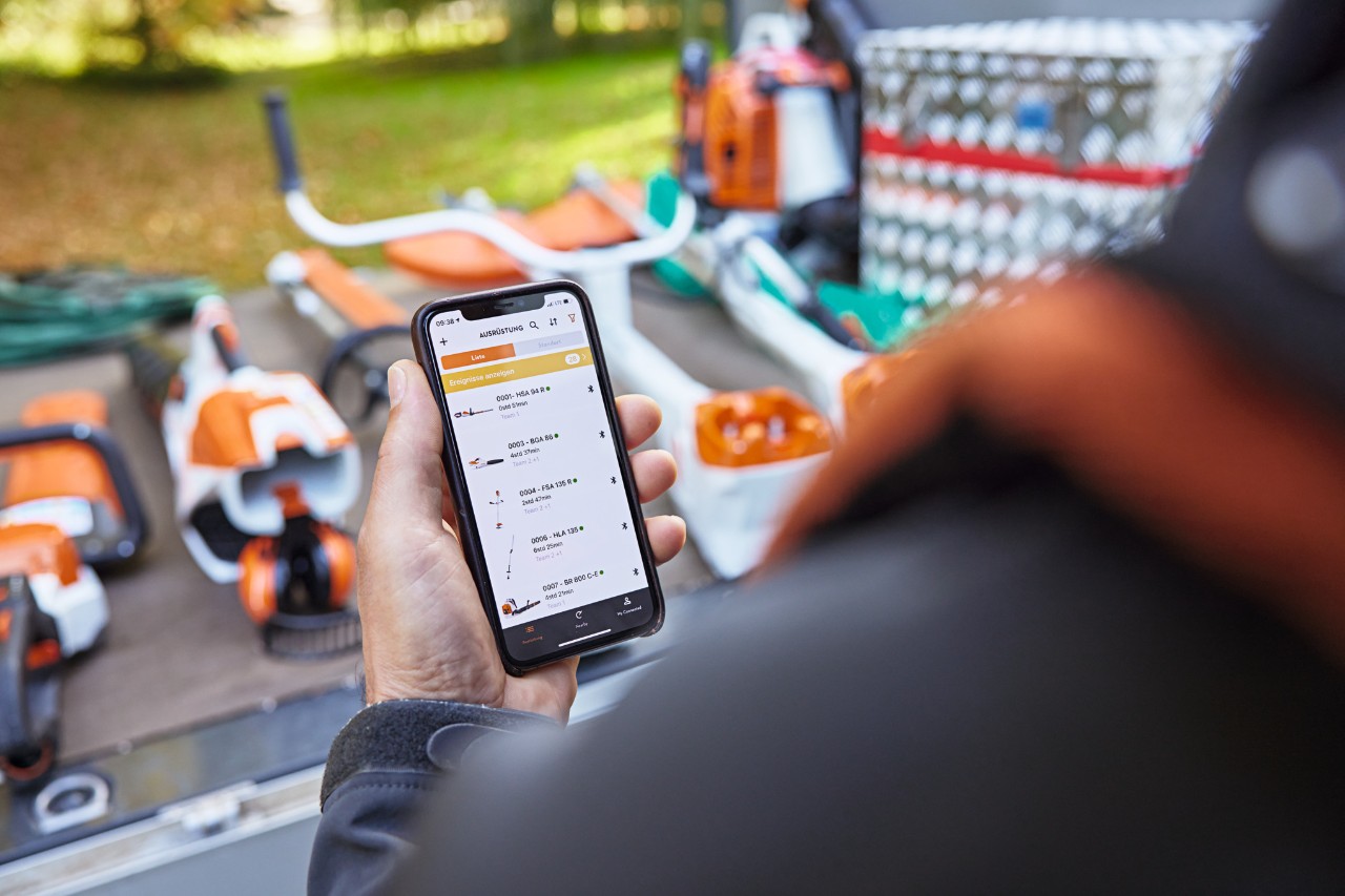 Osoba stojąca przed powierzchnią ładunkową ciężarówki z platformą, na której leżą różne urządzenia STIHL. Osoba trzyma w dłoni smartfon z otwartą aplikacją STIHL connected. 