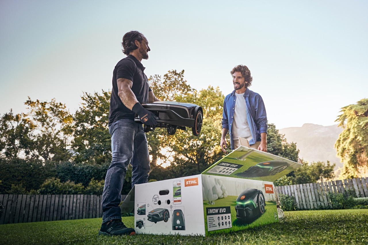 Dystrybutor STIHL trzyma w ręku iMOW® EVO, a karton iMOW® leży przed nim w ogrodzie klienta, któremu będzie pomagał w instalacji